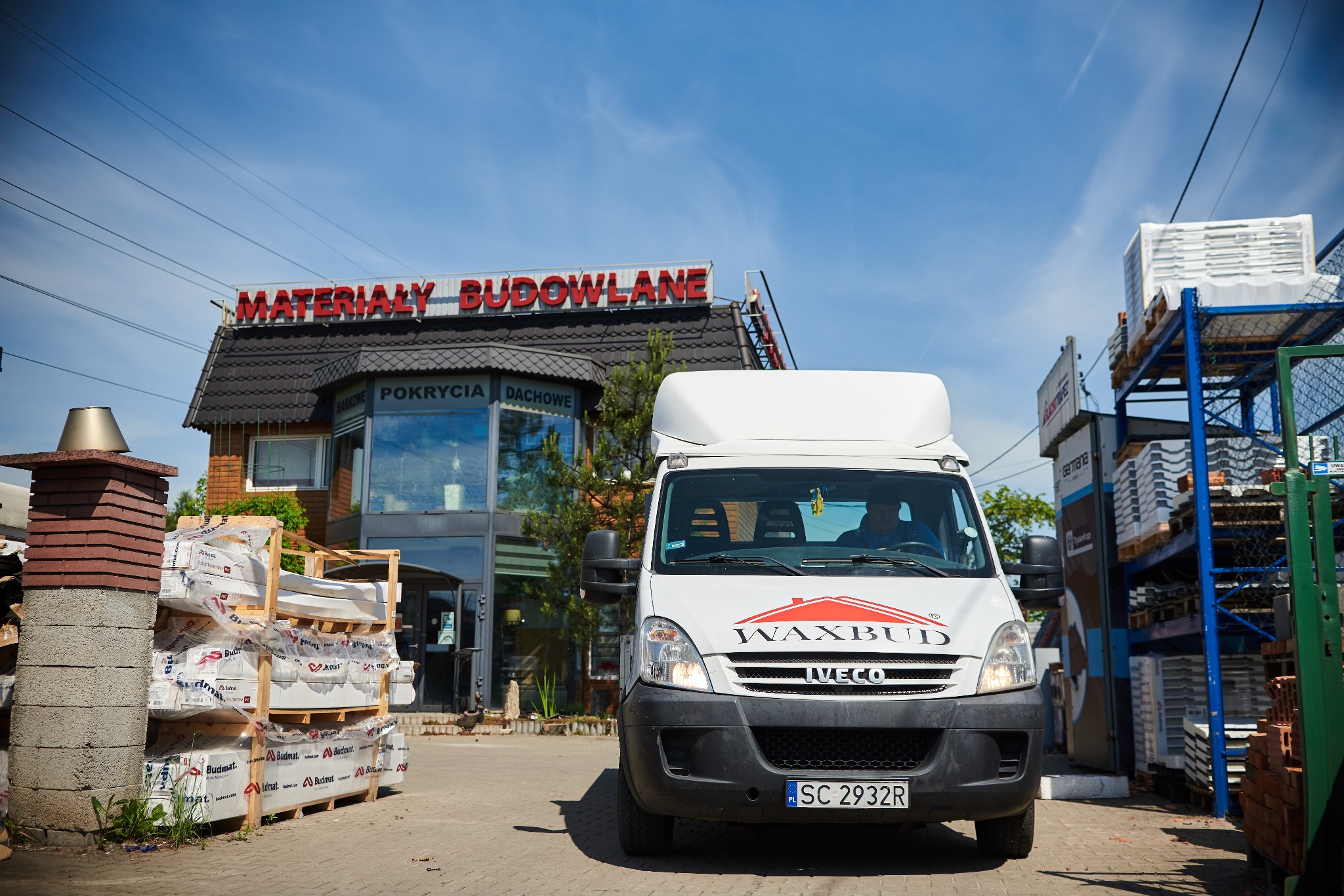 Biały samochód dostawczy firmy Waxbud Iveco na tle budynku sklepu z materiałami budowlanymi i pokryciami dachowymi, palety ze styropianem i dachówkami przed budynkiem.