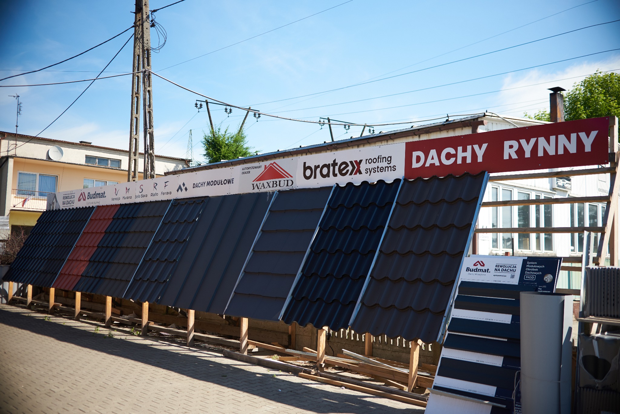 Ekspozycja różnych rodzajów pokryć dachowych, w tym blachodachówki i blachy trapezowej, prezentowanych na zewnątrz budynku z szyldami firm Budmat, Bratex i Waxbud, z widocznym fragmentem chodnika...
