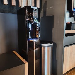 RAF-POLL PHU RAFAŁ ŚLIWA - Czarny dystrybutor wody Aqualogic z plastikowymi kubkami na blacie, obok kosz na śmieci ze stali nierdzewnej w biurowej przestrzeni z drewnianymi akcentami.