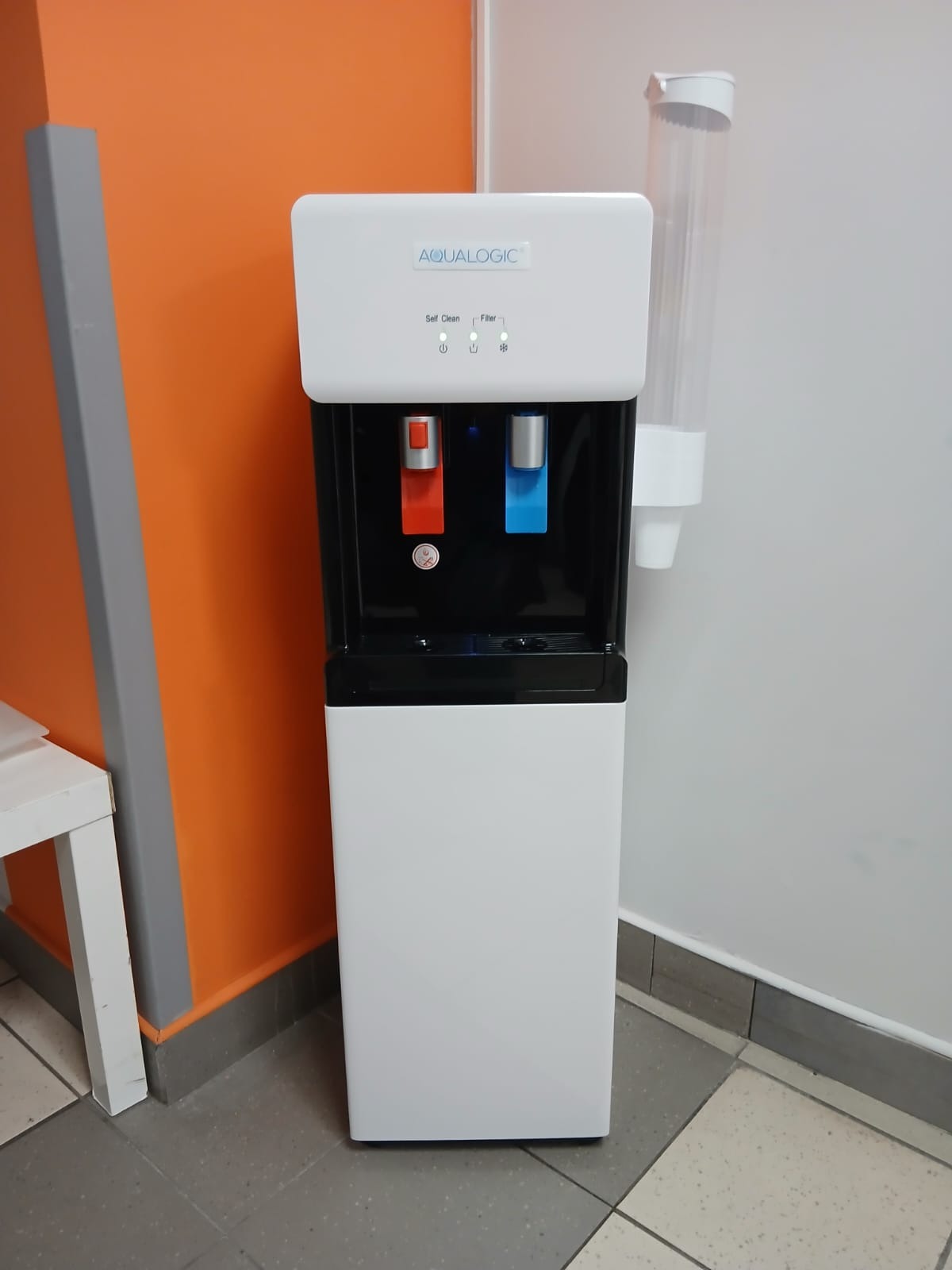 Nowoczesny, biały dystrybutor wody Aqualogic z dozownikami na ciepłą i zimną wodę, umieszczony w biurze z pomarańczową ścianą i dozownikiem na kubki.