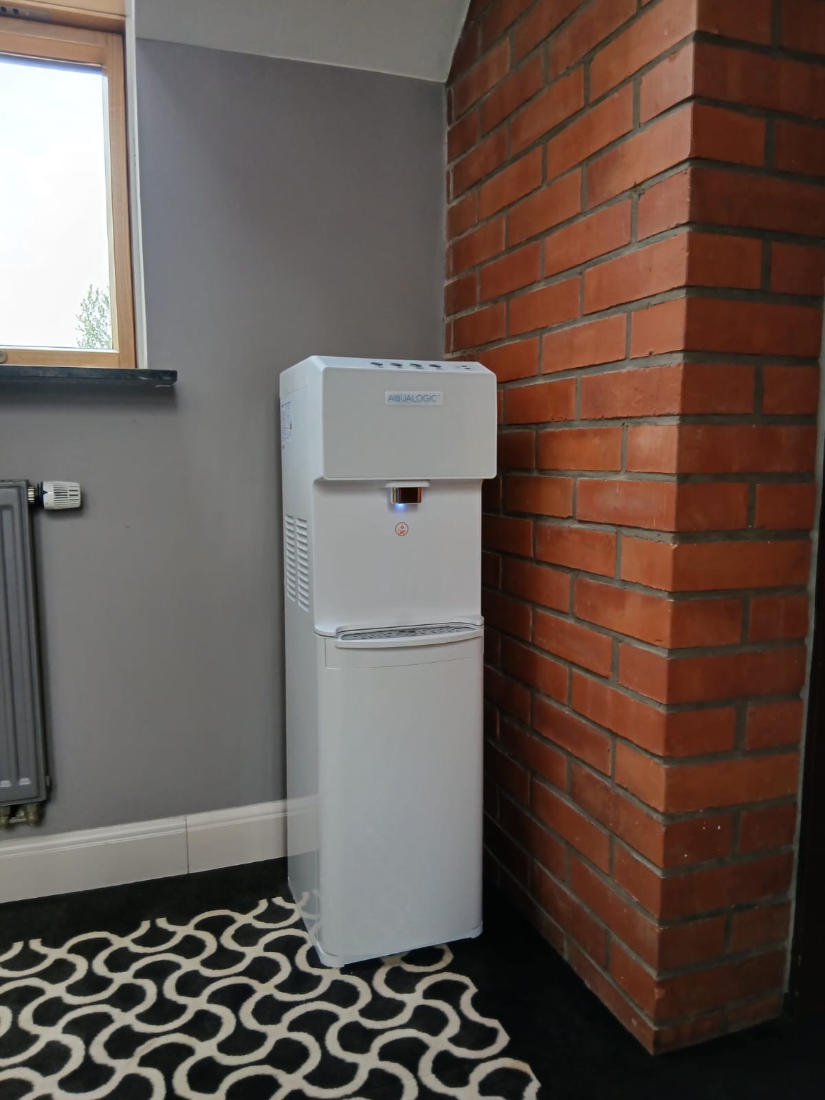 Biały dystrybutor wody Aqualogic stoi w rogu pomieszczenia, na tle szarej ściany i ceglanego filaru. Na podłodze wzorzysty dywan w czarno-białe fale.