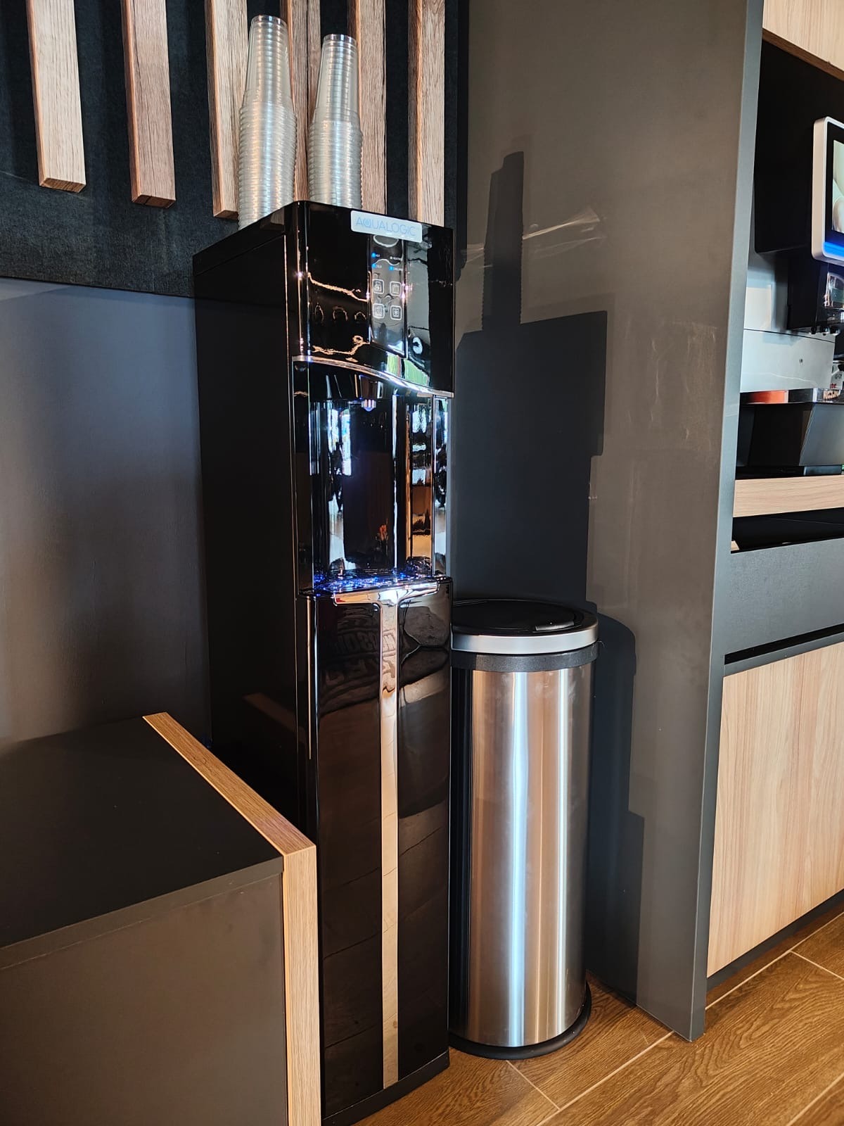 Czarny dystrybutor wody Aqualogic z plastikowymi kubkami na blacie, obok kosz na śmieci ze stali nierdzewnej w biurowej przestrzeni z drewnianymi akcentami.