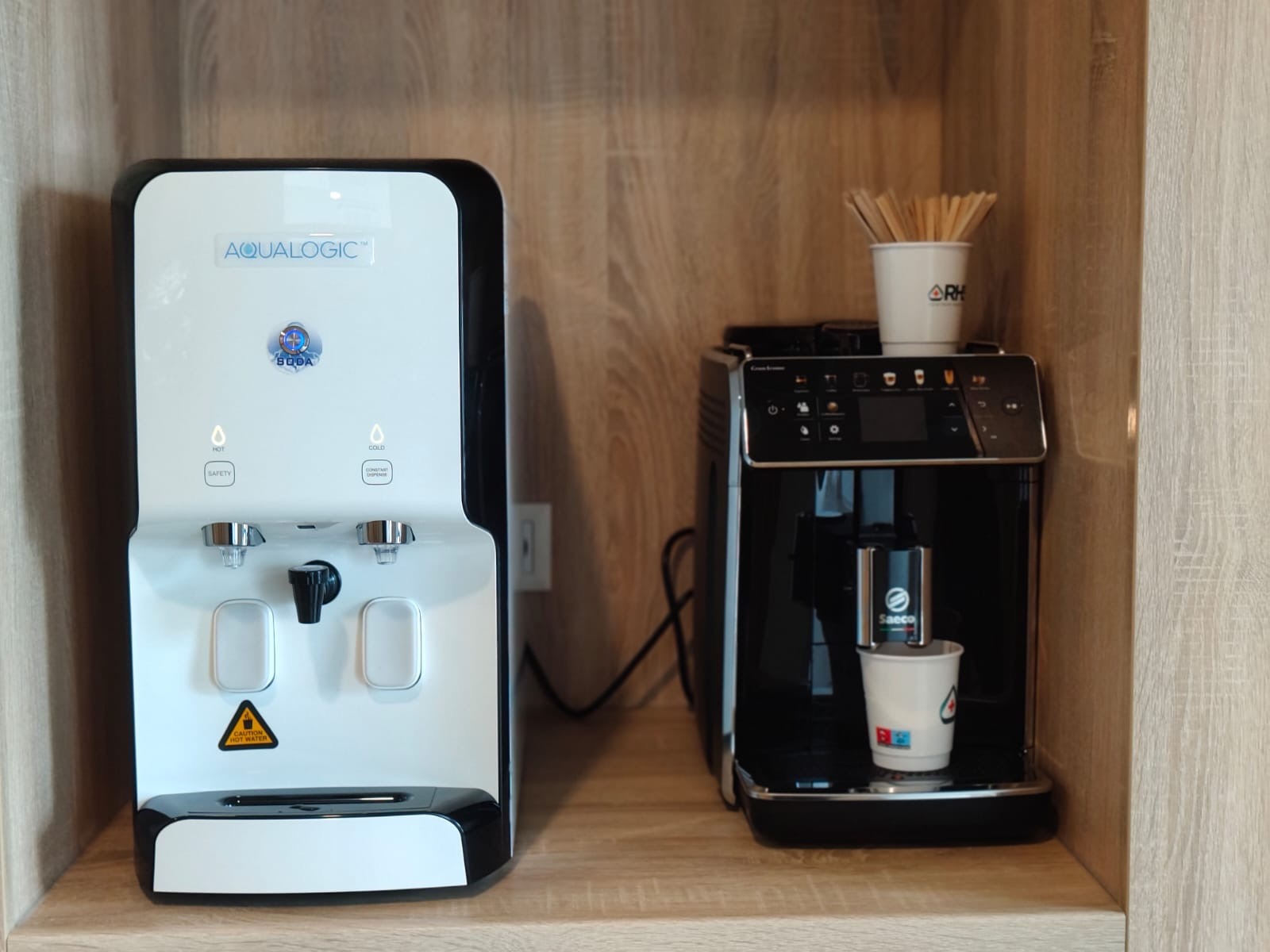 Dystrybutor wody Aqualogic obok ekspresu Saeco na drewnianym blacie. W kubku z ekspresu leje się kawa. Widok z przodu, oświetlenie dzienne.