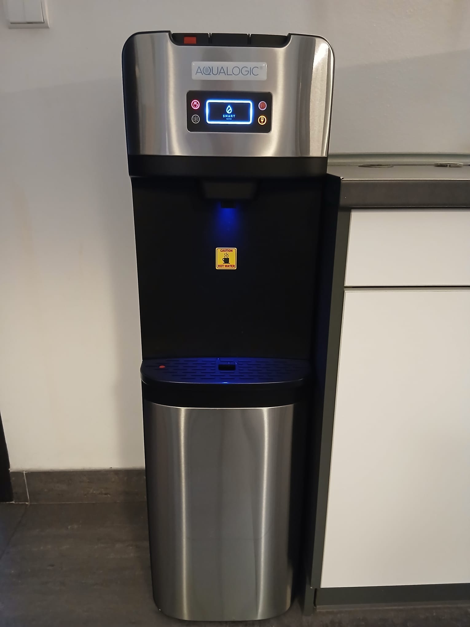 Nowoczesny dystrybutor wody Aqualogic ze stali nierdzewnej, z panelem dotykowym i funkcją gorącej wody, ustawiony w biurowej kuchni.