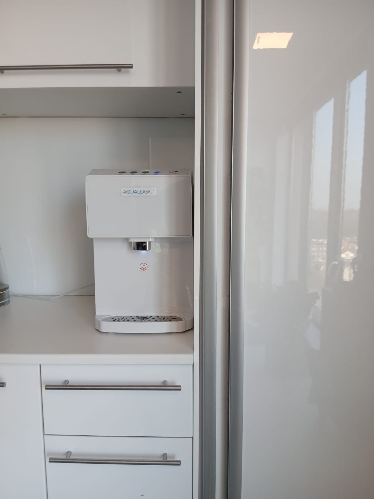 Biały dystrybutor wody Aqualogic na blacie kuchennym z białymi szafkami i srebrnymi uchwytami, obok błyszczącej, białej lodówki.