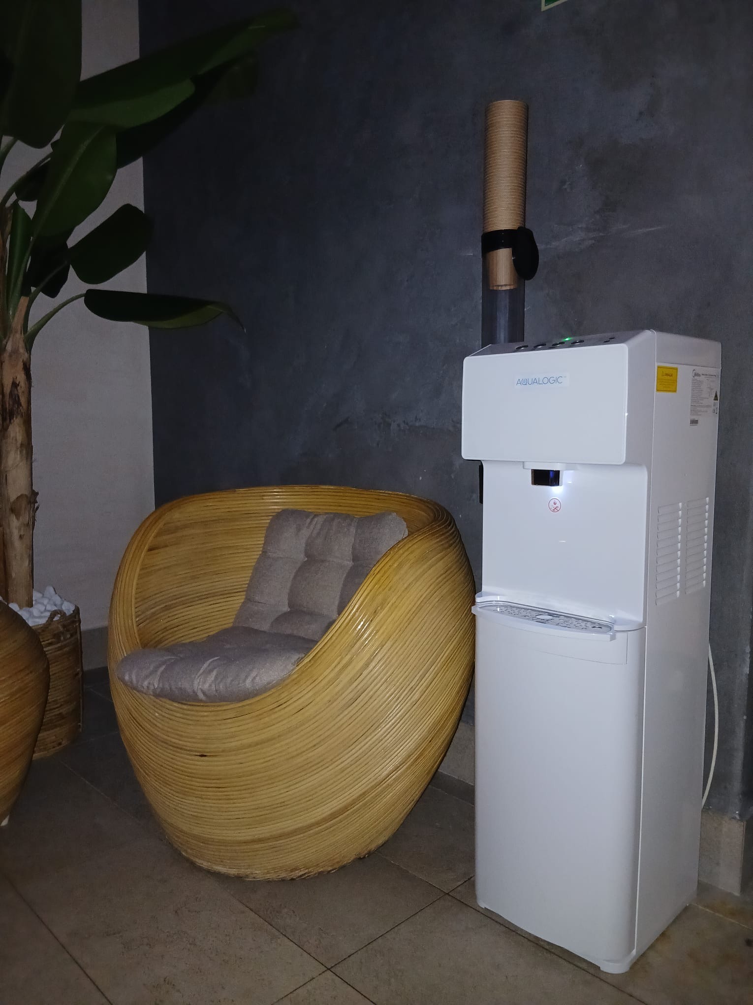 Biały dystrybutor wody Aqualogic obok fotela z rattanu i rośliny doniczkowej na tle szarej ściany. Nowoczesne wyposażenie biura lub poczekalni.