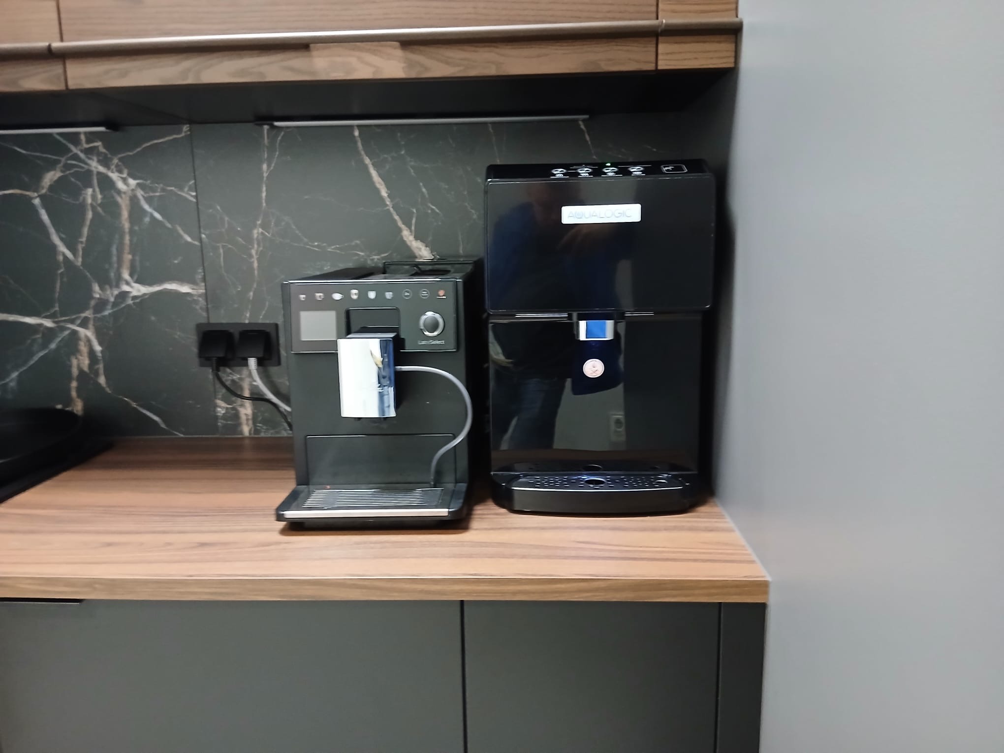 Czarny dystrybutor wody Aqualogic obok ekspresu do kawy na blacie kuchennym z marmurowym tłem. Nowoczesne rozwiązanie dla biura lub domu.