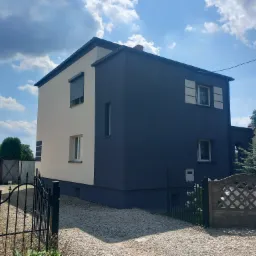 Dwukolorowa elewacja domu jednorodzinnego: kremowa i grafitowa, z widocznymi oknami i roletami, otoczona żwirowym podłożem i ogrodzeniem.