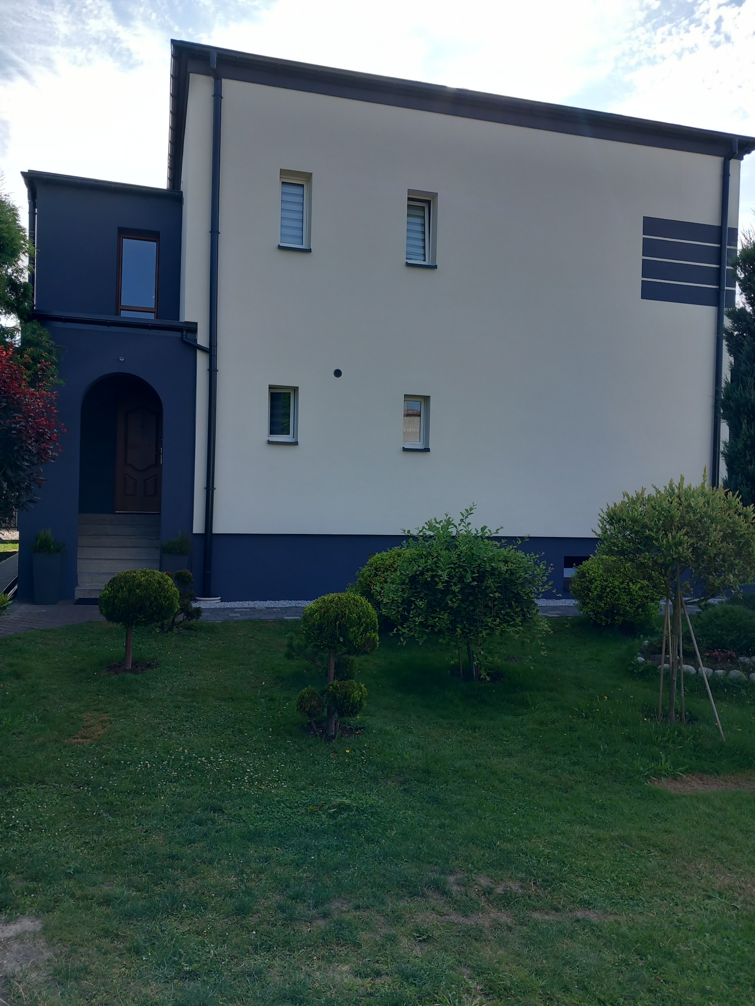 Dwukondygnacyjny dom jednorodzinny po renowacji elewacji, z kremową górną częścią i ciemnoszarą dolną częścią oraz wnęką wejściową, otoczony zadbanym trawnikiem i ozdobnymi krzewami.