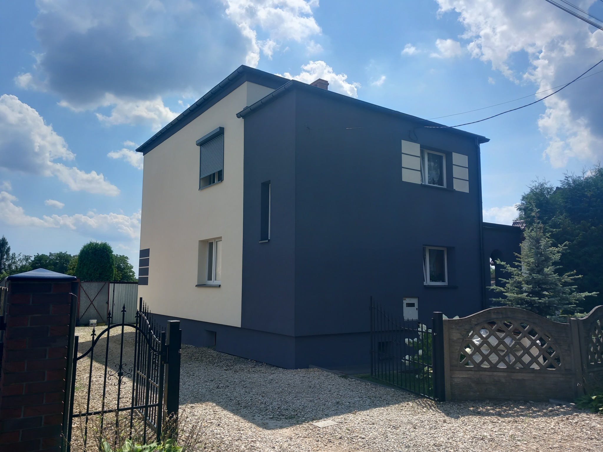 Dwukolorowa elewacja domu jednorodzinnego: kremowa i grafitowa, z widocznymi oknami i roletami, otoczona żwirowym podłożem i ogrodzeniem.