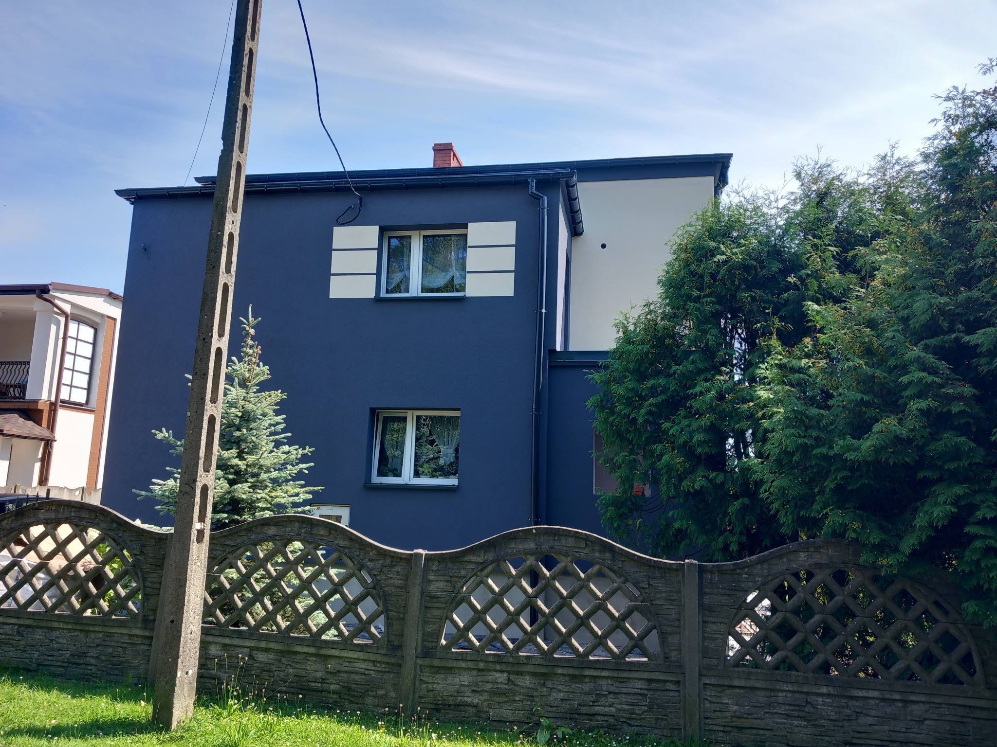 Dwukondygnacyjny dom z ciemnoszarą elewacją, białymi akcentami wokół okien i jasną ścianą boczną, widziany zza betonowego płotu o ażurowym wzorze, z zielenią drzew i krzewów w tle oraz słupem...