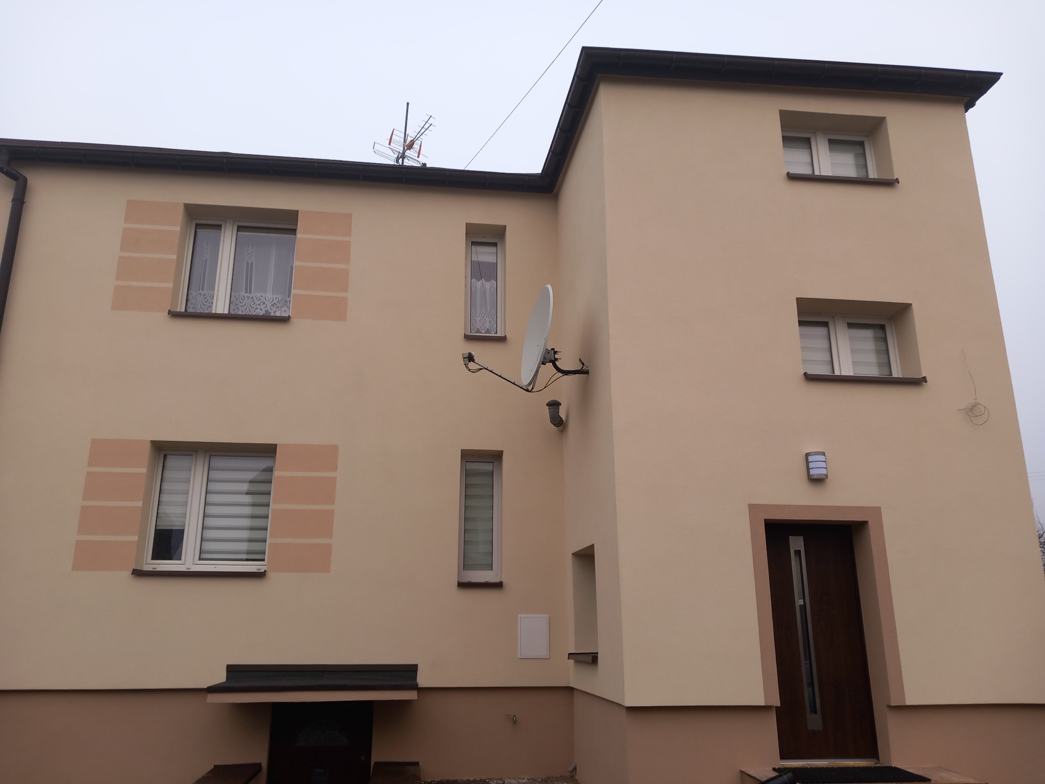 Ocieplony budynek jednorodzinny z beżową elewacją, widoczne okna z dekoracyjnymi obramowaniami, antena satelitarna i lampa zewnętrzna przy wejściu.