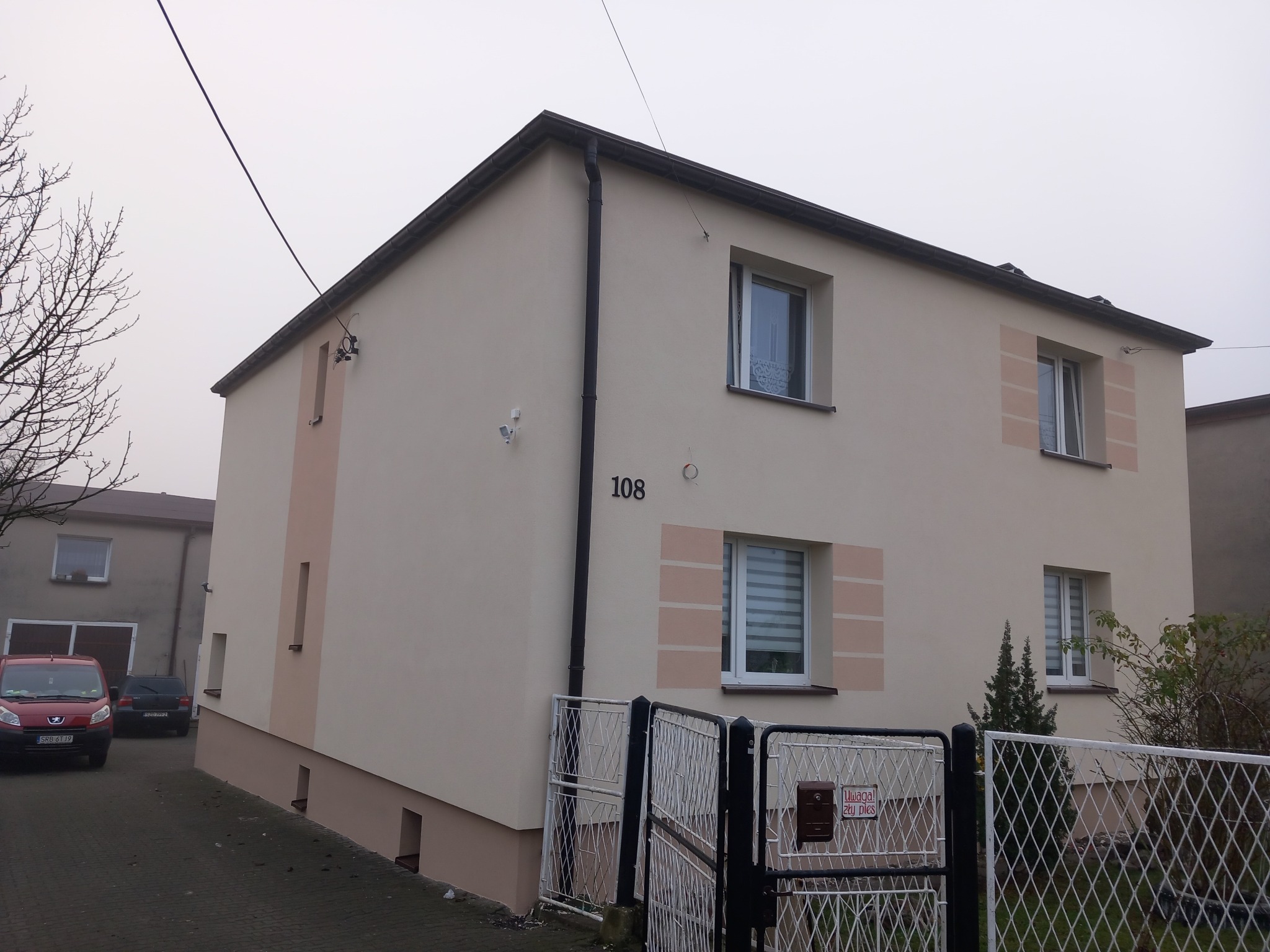 Dwupiętrowy dom z beżową elewacją i dekoracyjnymi, różowymi elementami wokół okien, widoczny numer domu '108' i fragment ogrodzenia z tabliczką ostrzegawczą.