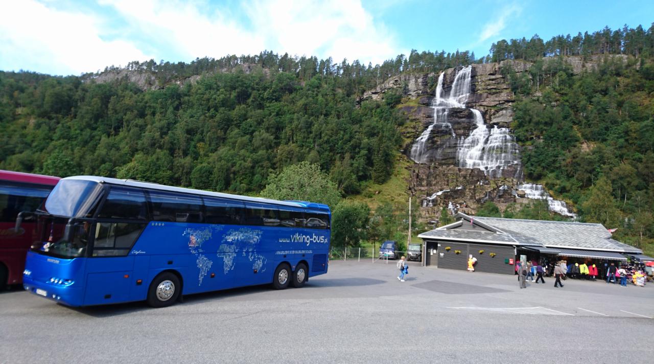 Niebieski autokar turystyczny z mapą świata i napisem 'www.viking-bus.pl' na parkingu z widokiem na wodospad i budynek z pamiątkami.