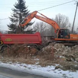 Pomarańczowa koparka Doosan ładuje gruz do czerwonej wywrotki Meller na tle zimowego krajobrazu z drzewami i śniegiem przy drodze.