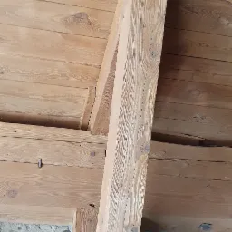 Drewniany strop z elementami konstrukcyjnymi z belek, widoczna surowa cegła na ścianie w tle.