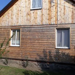 Drewniana elewacja domu przed i po czyszczeniu, widoczny kontrast między górną, jaśniejszą częścią a dolną, ciemniejszą, brudną częścią, trzy okna z białymi ramami.