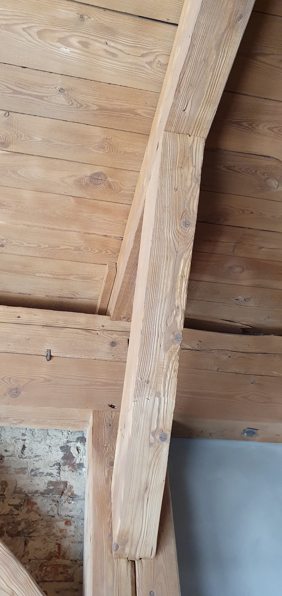 Drewniany strop z elementami konstrukcyjnymi z belek, widoczna surowa cegła na ścianie w tle.