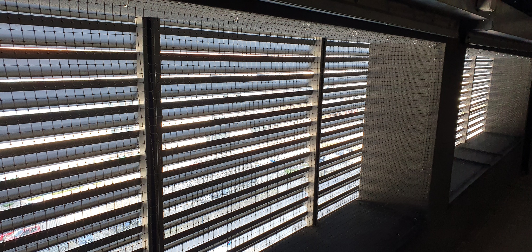 Balkon zabezpieczony siatką ochronną przed ptakami, widoczne żaluzje i fragmenty zabudowy balkonu.