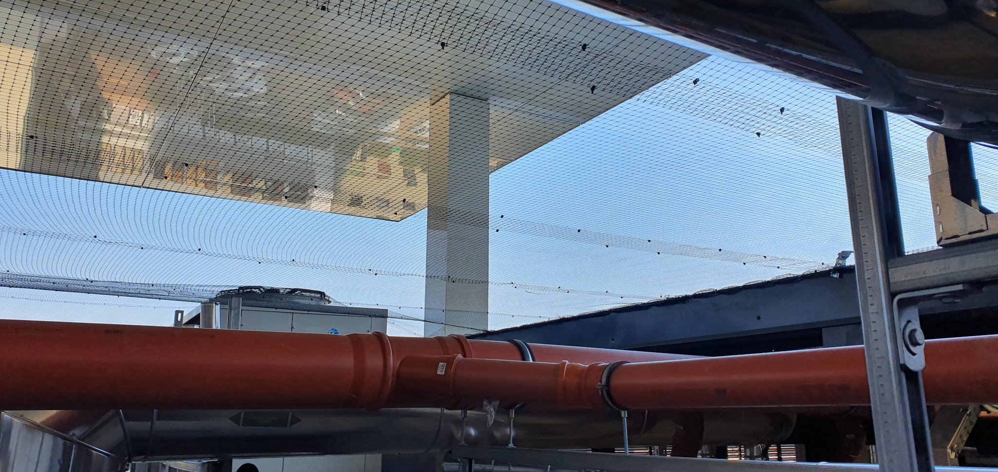 Ochronna siatka przeciw ptakom zamontowana pod zadaszeniem, widoczne pomarańczowe rury instalacyjne i fragmenty systemów wentylacyjnych.