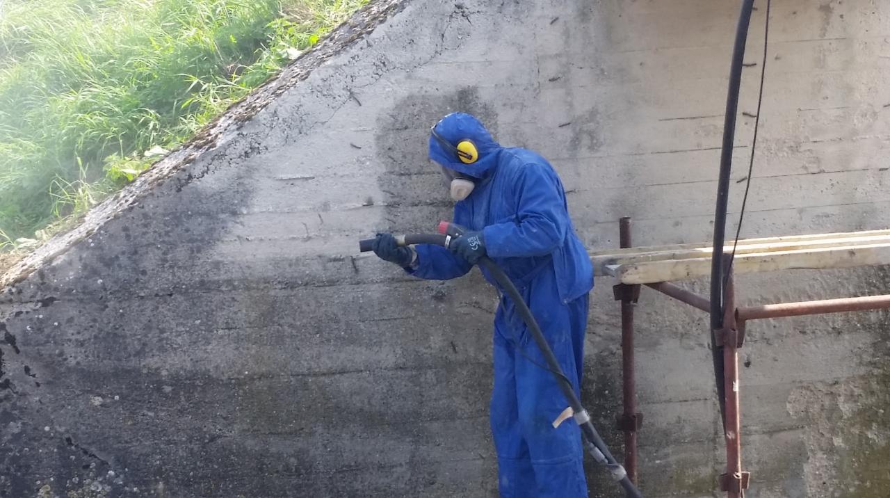 Pracownik w kombinezonie ochronnym i masce oczyszcza betonową ścianę z zabrudzeń i mchu za pomocą specjalistycznego sprzętu. Widoczny fragment rusztowania i otoczenie z zielenią.