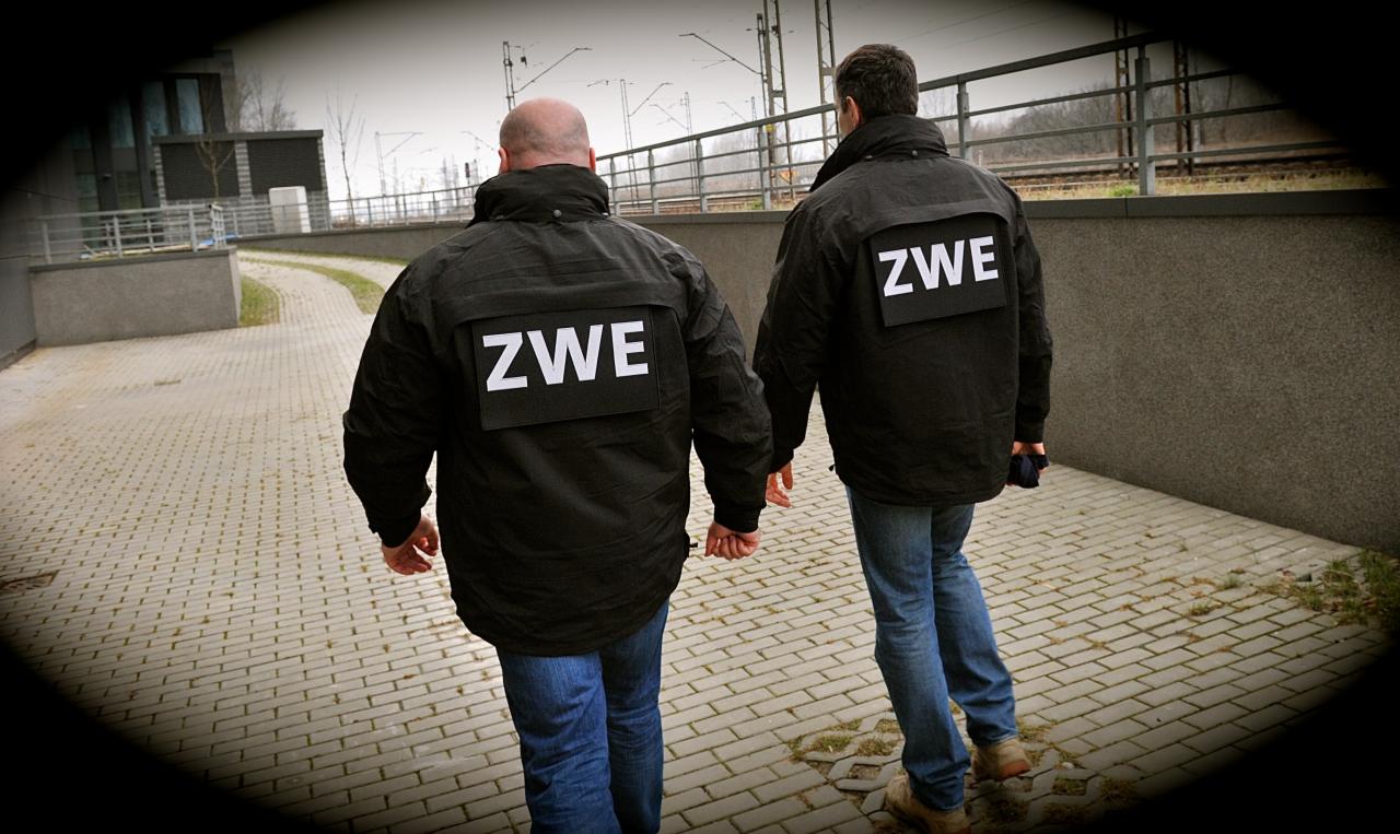 Dwóch mężczyzn w czarnych kurtkach z napisem 'ZWE' na plecach idzie po chodniku. W tle widoczne tory kolejowe i budynek.