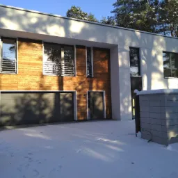 Nowoczesny dom z elewacją z drewnianych paneli i szarą bramą garażową w zimowej scenerii, śnieg na podjeździe.