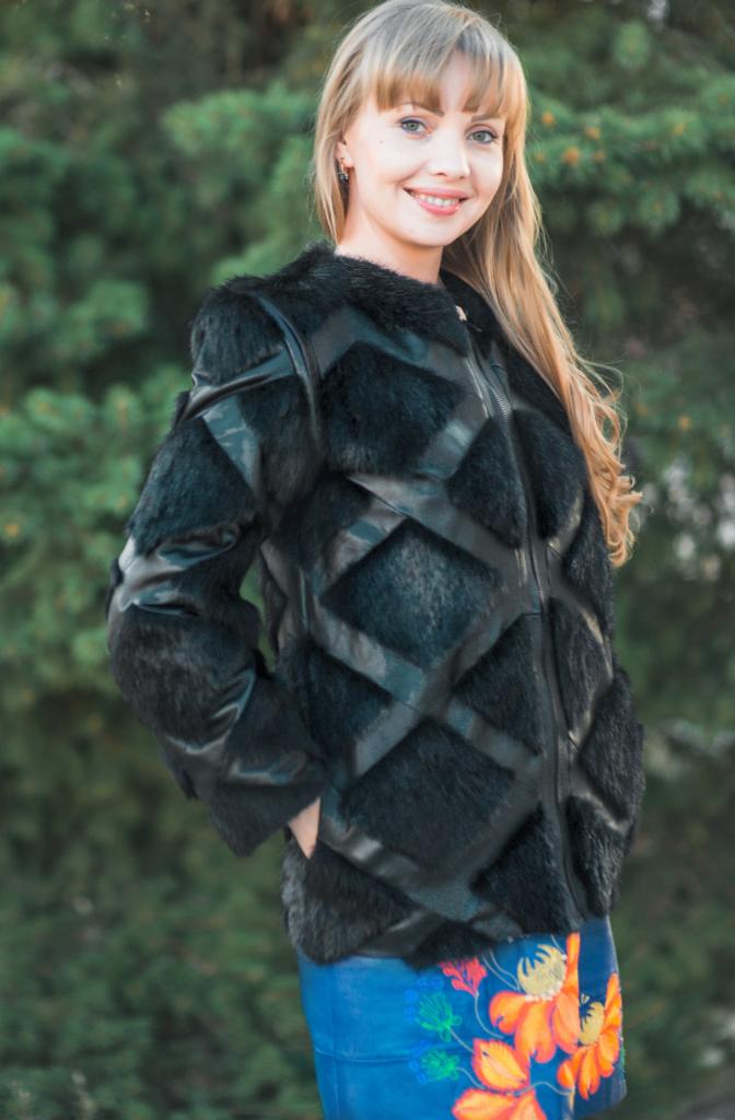 Uśmiechnięta blondynka w czarnej kurtce z geometrycznym wzorem z futra i skóry, na tle zieleni.