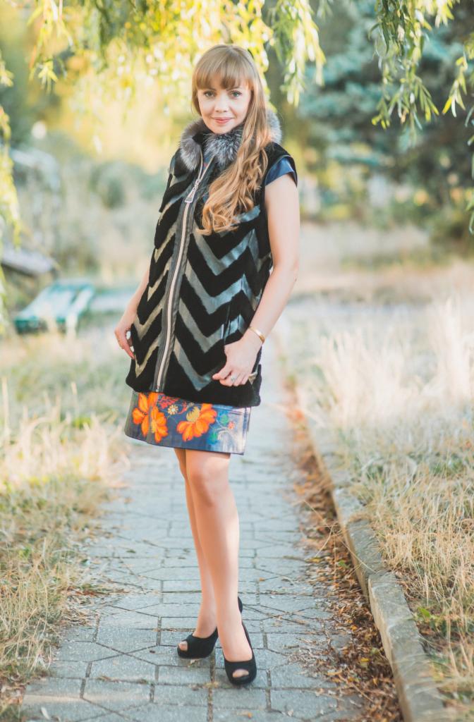 Kobieta w czarnej kamizelce ze wzorem w zygzaki i futrzanym kołnierzem, pozująca na ścieżce w parku, z widoczną kwiecistą spódnicą i czarnymi butami na obcasie.