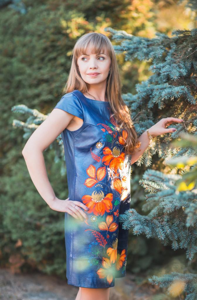 Kobieta w niebieskiej sukience z pomarańczowymi haftowanymi kwiatami, pozująca na tle zielonych drzew.