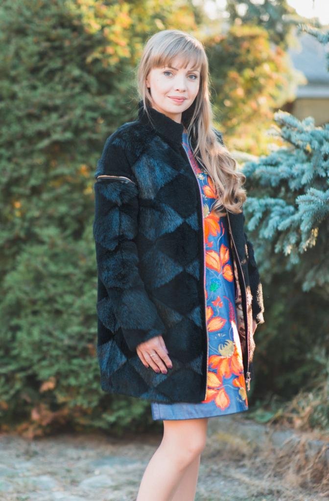 Kobieta w czarnym futrze z geometrycznym wzorem rombów, zamek błyskawiczny delikatnie rozpięty, ukazujący kolorową sukienkę w kwiaty, na tle zieleni.