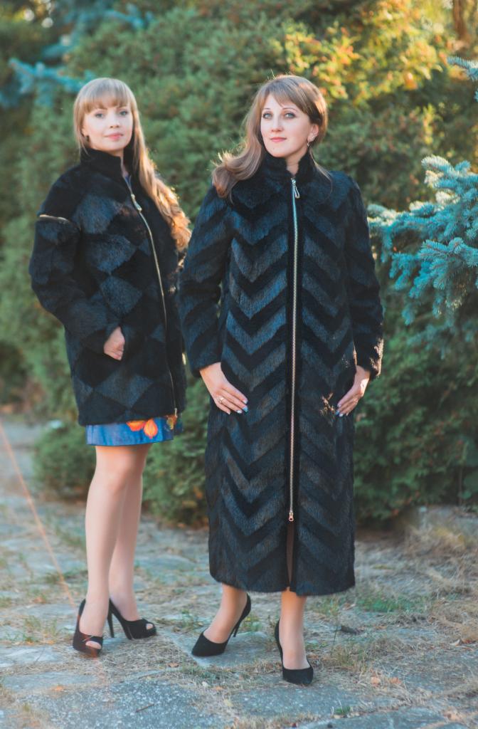 Dwie kobiety prezentują czarne, futrzane płaszcze z geometrycznym wzorem i zamkiem błyskawicznym na tle zielonych krzewów.