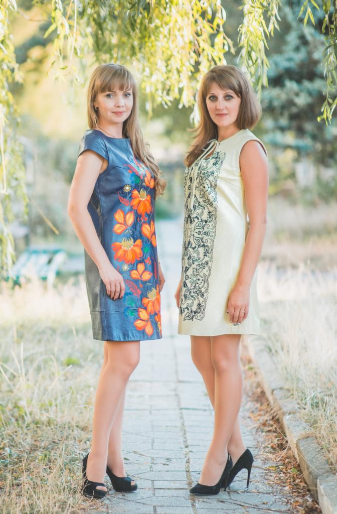 Dwie kobiety prezentują sukienki: granatowa z malowanymi pomarańczowymi kwiatami i kremowa z haftowanym czarnym wzorem, na tle alei parkowej.