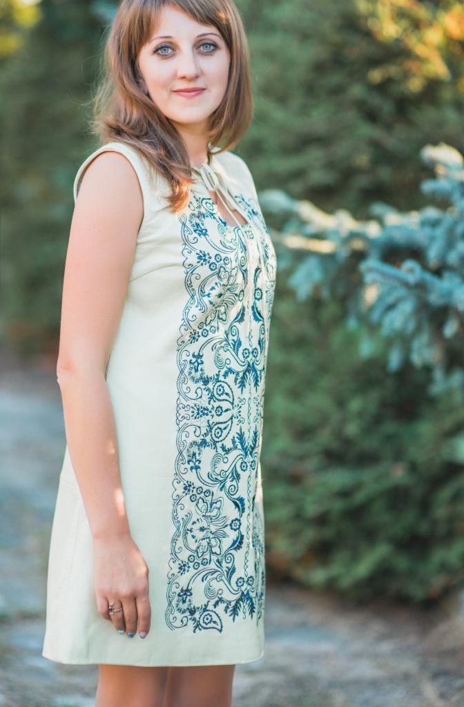 Kobieta w kremowej sukience z niebieskim haftem kwiatowym, pozująca na tle zieleni.