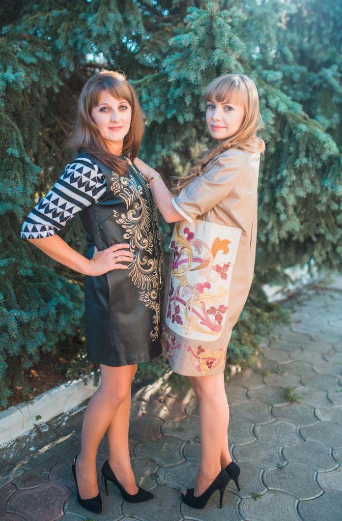 Dwie kobiety prezentują autorskie sukienki: czarna, skórzana z haftowanymi złotymi ornamentami i beżowa z aplikacjami kwiatowymi, na tle zielonego drzewa.