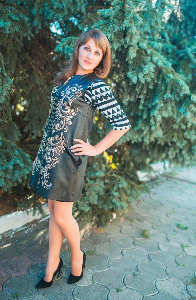 Kobieta prezentuje czarną sukienkę z haftowanymi, złotymi wzorami i geometrycznym motywem na rękawach, pozując na tle zielonych drzew.