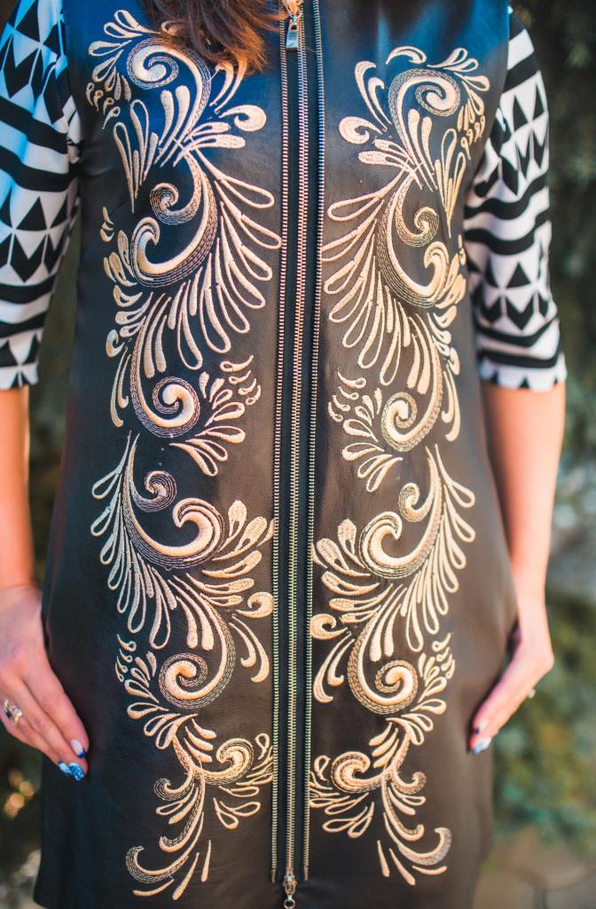 Czarna sukienka z ekoskóry z haftowanymi, symetrycznymi zdobieniami w kolorze ecru, zamek błyskawiczny na całej długości, rękawy w biało-czarne geometryczne wzory.