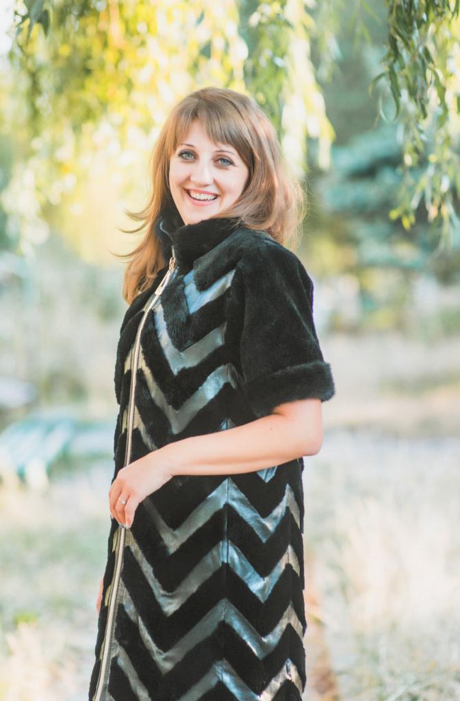 Uśmiechnięta kobieta w czarnym płaszczu z zygzakowatym wzorem i złotym zamkiem błyskawicznym, na tle zieleni.