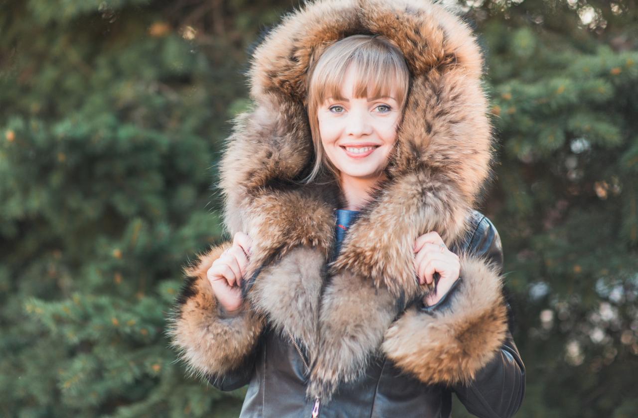 Uśmiechnięta blondynka w skórzanej kurtce z futrzanym kapturem i mankietami na tle zielonych drzew iglastych.