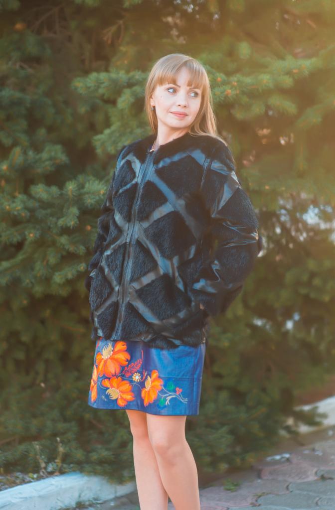 Kobieta w czarnej kurtce z futrzanymi wstawkami i niebieskiej spódnicy z haftowanymi kwiatami, pozująca na tle zielonego drzewa.  Ujęcie z delikatnym efektem bokeh.