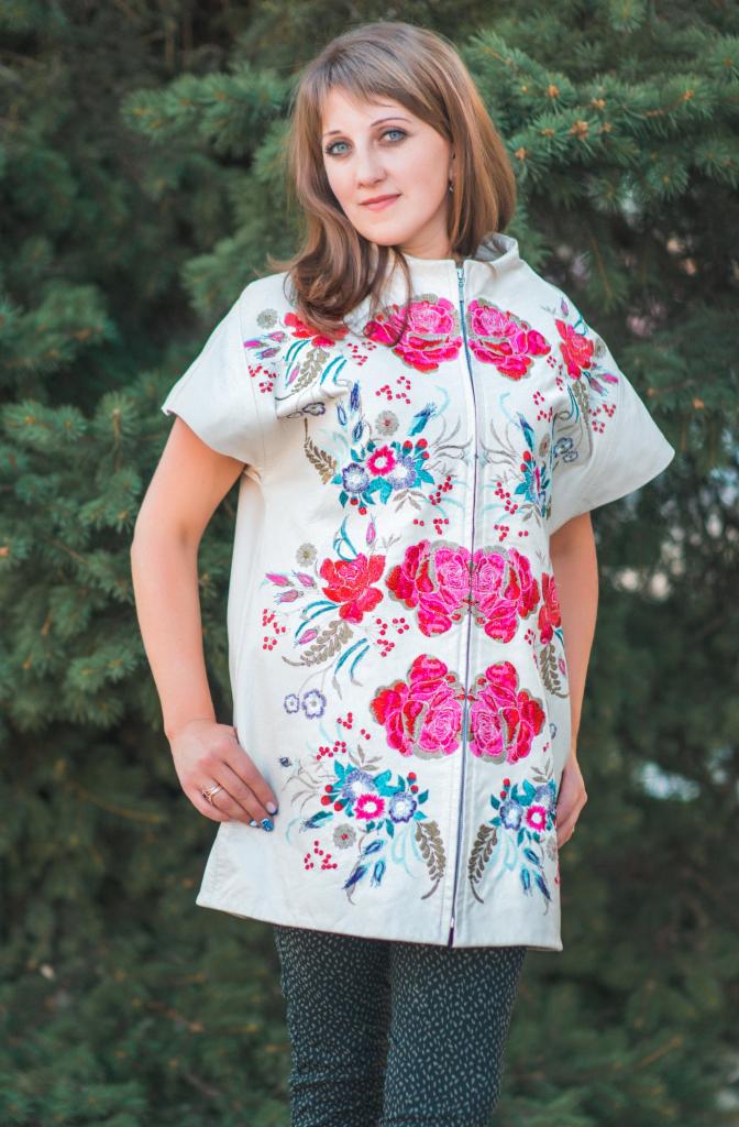 Kobieta prezentuje białą kamizelkę zapinaną na zamek, ozdobioną bogatym, kolorowym haftem kwiatowym, na tle zielonego iglastego drzewa.