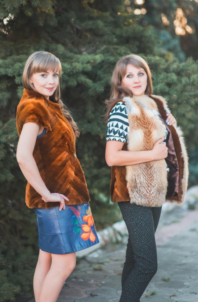 Dwie kobiety prezentują futrzane kamizelki: jedna w brązowej, krótko strzyżonej kamizelce z podszewką w kolorze burgundu i niebieskiej spódnicy z kwiatowym wzorem, druga w kamizelce z długiego...