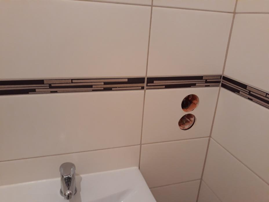 Białe płytki ścienne z dekoracyjnym pasem i dwoma otworami przygotowanymi pod instalację baterii umywalkowej, widoczna umywalka z baterią.