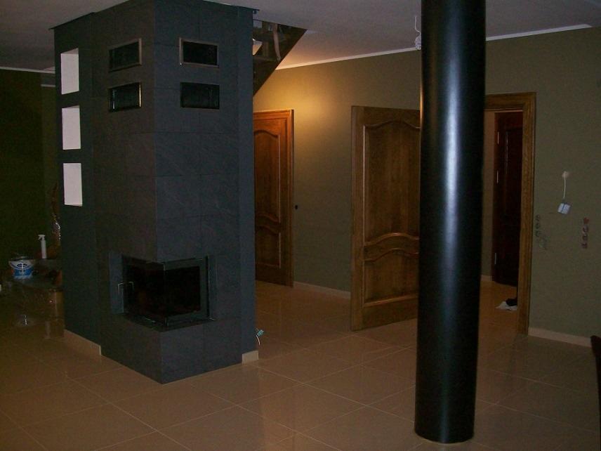 Obudowa kominka z ciemnych, kwadratowych płytek, z otworami dekoracyjnymi, na podłodze z płytek w odcieniach brązu, widoczne trzy drewniane drzwi w tle.