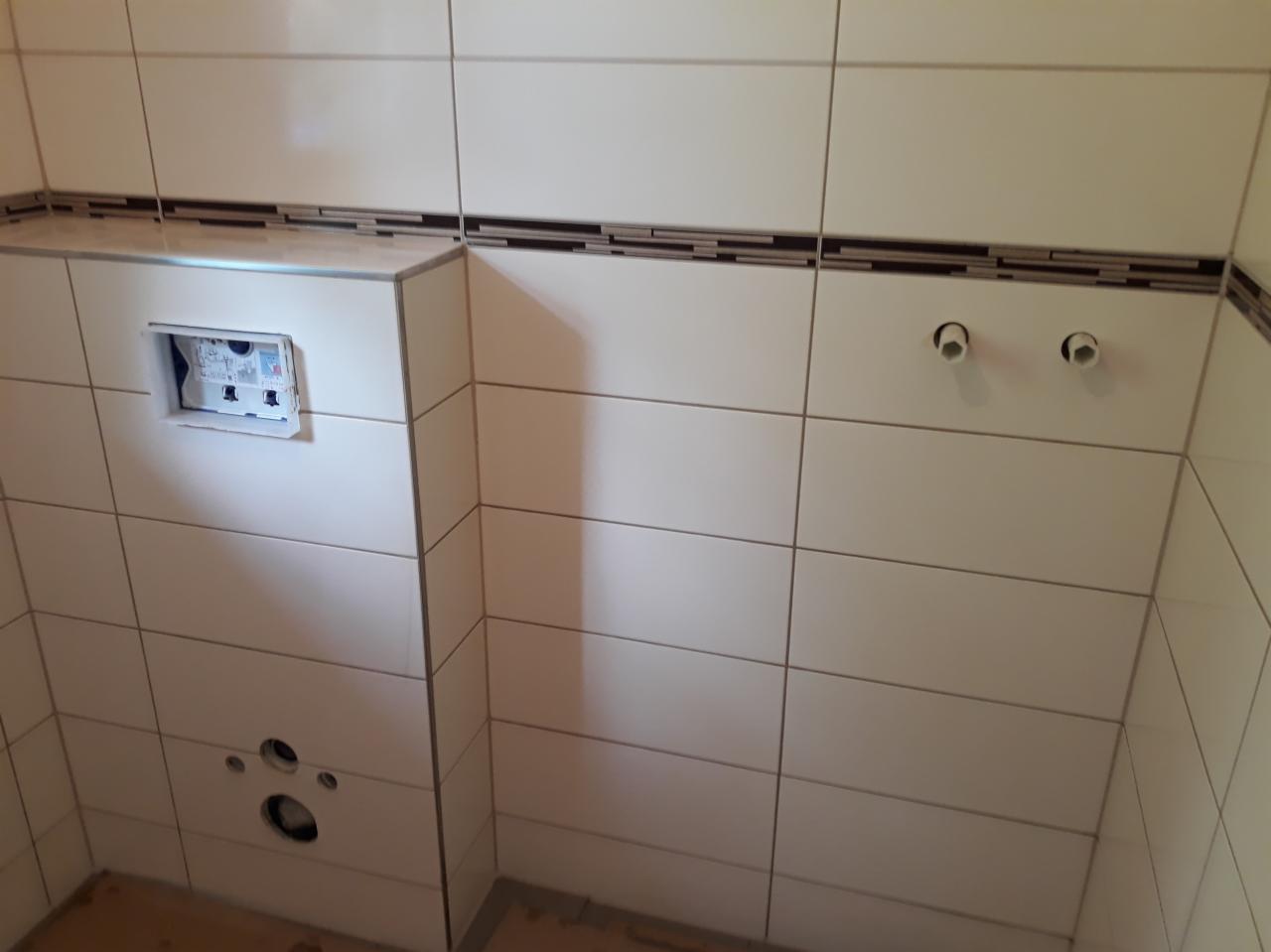 Białe, błyszczące płytki ceramiczne ułożone na ścianach łazienki, z dekoracyjnym paskiem mozaiki w odcieniach brązu, widoczne otwory na instalacje sanitarne i przygotowane miejsce na stelaż...