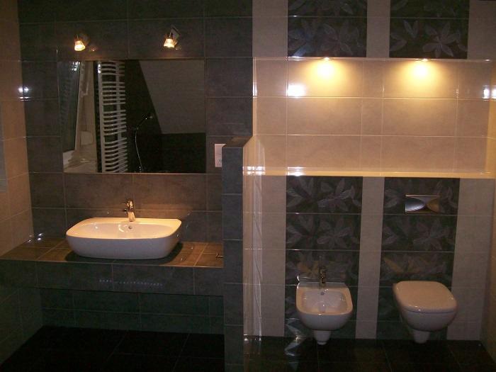 Wykończona łazienka z szarymi i beżowymi płytkami z motywem liści, białą umywalką nablatową, bidetem i toaletą wiszącą, oświetlona punktowymi lampami.