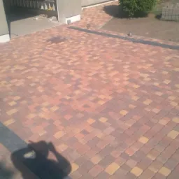 Ułożona kostka brukowa w odcieniach brązu i pomarańczy z czarnymi obrzeżami, prowadząca do garażu i ogrodu, widoczny cień fotografa.