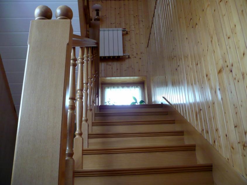 Drewniane schody z balustradą i toczonymi tralkami, widok od dołu. Ściany wyłożone boazerią, w tle okno z firanką i grzejnik.