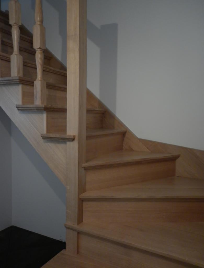 Drewniane schody z balustradą w naturalnym odcieniu drewna, widoczne z bliska, z elementami toczonymi i prostymi. Minimalistyczny design, jasne drewno, neutralne tło.