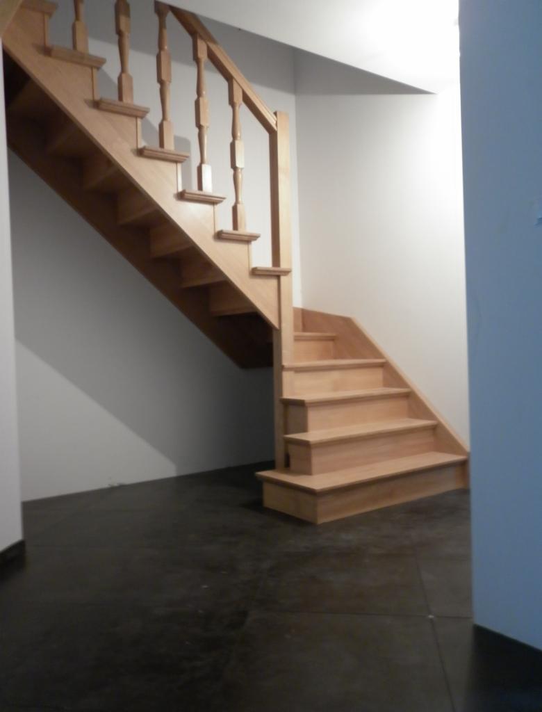 Jasne, drewniane schody z balustradą w minimalistycznym wnętrzu. Ciemna posadzka kontrastuje z bielą ścian. Widoczne detale wykończenia schodów.